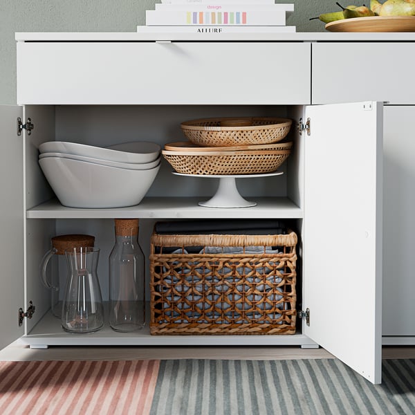 Armoire blanche avec deux étagères, bols, plateaux, bocaux et panier, tapis gris en dessous.