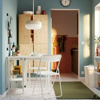 Cuisine moderne avec armoires en bois blanc et murs bleus. Chaise pliante VIHALS blanche contre table en pin. Design lumineux, spacieux et simple.