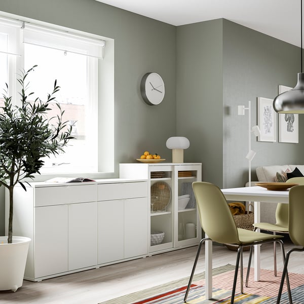 Salle à manger moderne avec rangement blanc VIHALS, portes vitrées et chaises vert pâle à côté d’une fenêtre.