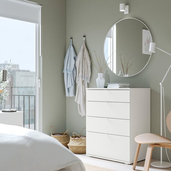 Chambre moderne avec mur vert, commode blanche, miroir rond et chaise en bois. Les vêtements sont suspendus à des crochets, les plantes ajoutent de la nature.