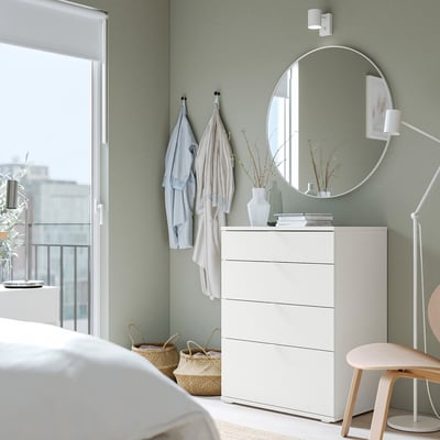 Chambre moderne avec mur vert, commode blanche, miroir rond et chaise en bois. Les vêtements sont suspendus à des crochets, les plantes ajoutent de la nature.