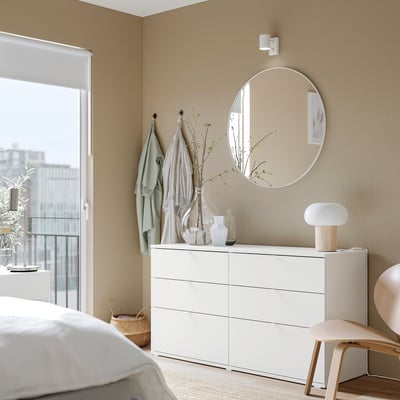 Une chambre minimaliste avec une commode VISALS blanche. Il y a six tiroirs, un miroir rond et un décor de couleur claire.