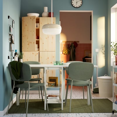 Salle à manger moderne avec table VIHALS blanche et chaises vertes. Des armoires en bois clair, des murs bleus et une cloison de séparation visible en arrière-plan.