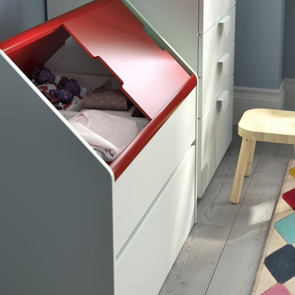 Coffre à jouets ouvert à couvercle rouge avec des couvertures et des animaux en peluche, un tiroir gris et un petit tabouret en bois.