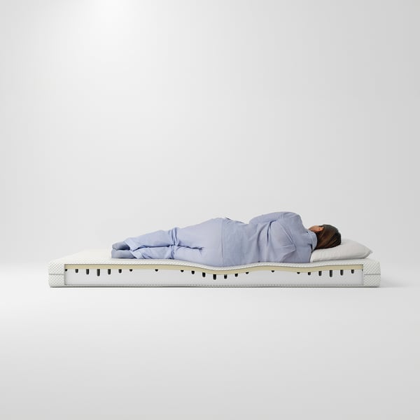 Persoon slaapt op een lichtblauwe ÅBYGDA-matras, die 16 cm hoog is met comfortzones, tegen een witte achtergrond.