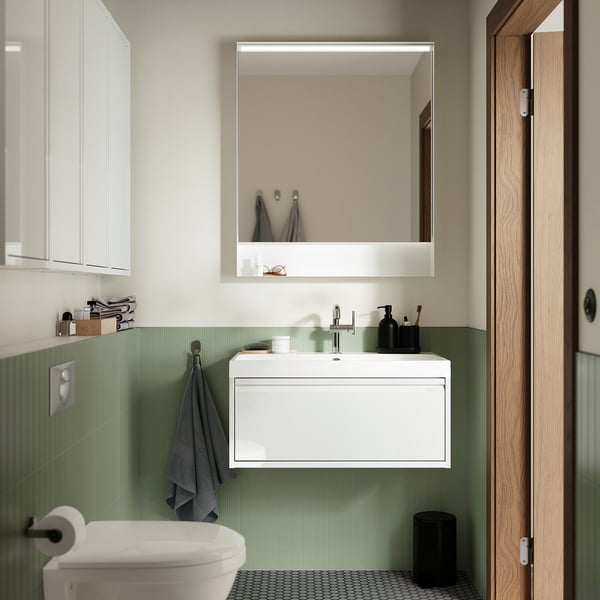 Moderne badkamer met mintkleurige tegels, een aan de muur gemonteerde witte wastafelkast, vierkante spiegel en zwarte accenten. Een grijze handdoek hangt naast het toilet.