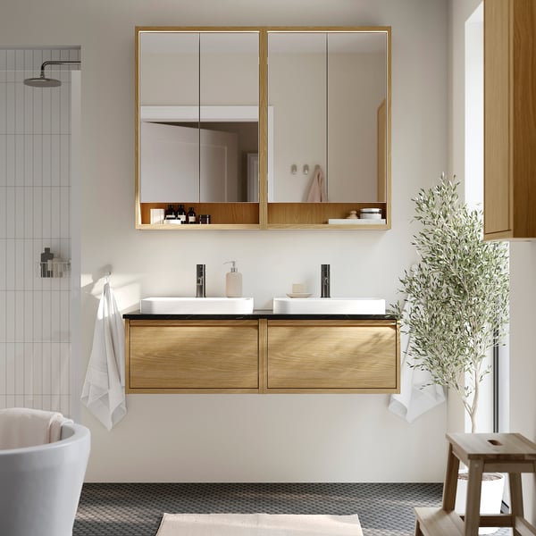 Moderne badkamer met een eiken dubbele wastafel, spiegelkast, witte armaturen. Decoratieve plant en kruk bij het raam. Handdoeken per bad.