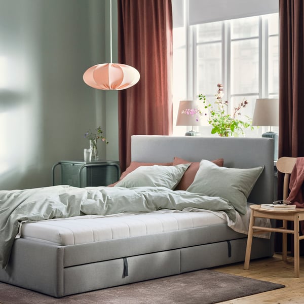 Gezellige slaapkamer met grijze ÅFJÄLL matras op houten vloer bij raam. Zachte verlichting van roze lamp, groene muren en roze gordijnen.