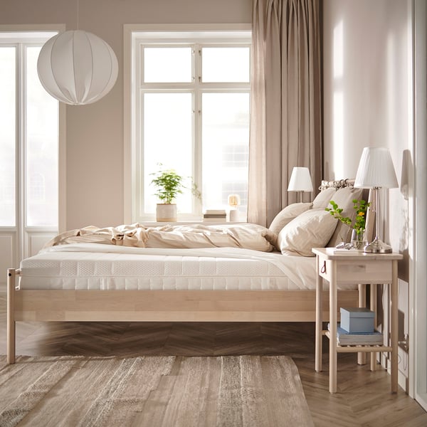 Slaapkamer met ÅKREHAMN-bed , linnenbed, houten vloer, gordijnen, hanglamp, bijzettafeltjes, tapijt en plant.
