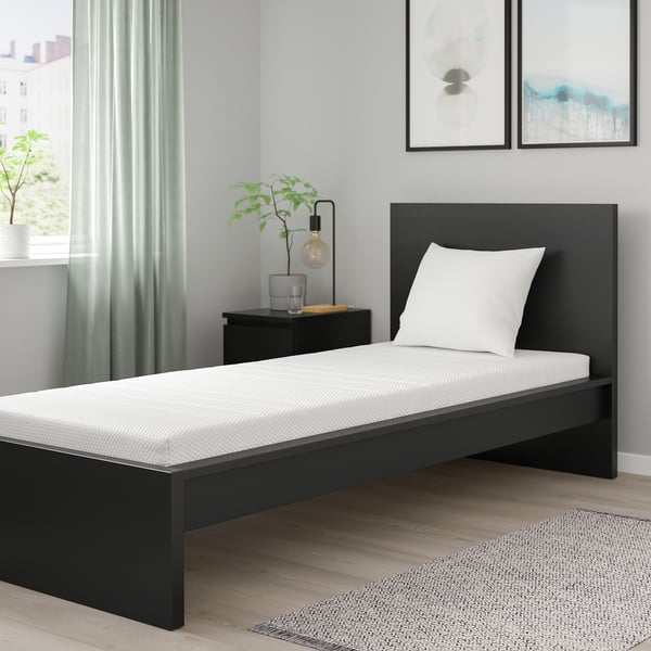 Moderne slaapkamer met zwart bed, wit kussen, grijze muur, nachtkastje, lamp, plant, frames, lichte houten vloer, vloerkleed.