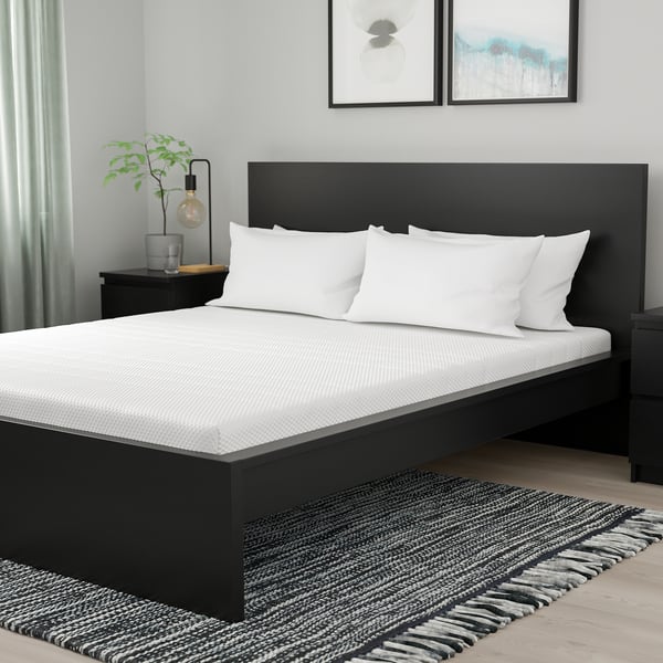 Moderne slaapkamer met zwarte queensize matras, witte kussens, nachtkastje, lamp, plant, kunst en gestreept vloerkleed.