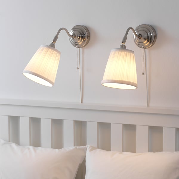 Twee aan de muur gemonteerde lampen met beige kappen die naar beneden zijn gericht, verlichten een wit bed met kussens. Trekkoorden voor schakelaars zichtbaar.