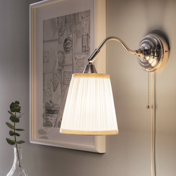 Een wandlamp met een verstelbare hals en een witte geplooide kap straalt zacht licht uit naast een ingelijste foto.