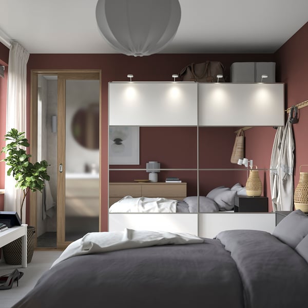 Slaapkamer met rode muren, witte schuifkast, grijs beddengoed, witte kussens, hangende jassen en planten.
