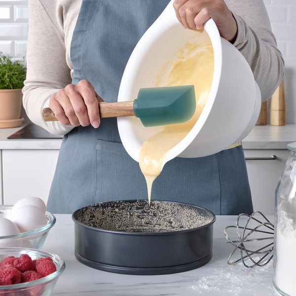 Een persoon giet beslag uit een witte kom met behulp van een groene spatel in een ronde pan, omgeven door bakingrediënten op een aanrecht.