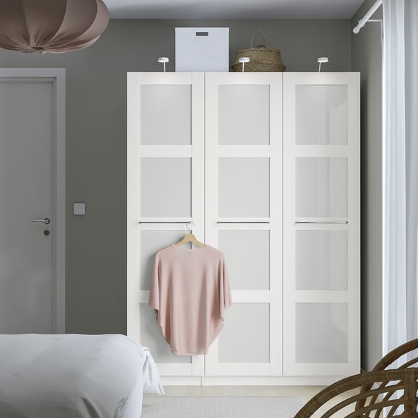 Minimalistische witte BERGSBO kledingkast met matglas, hangend blush shirt, naast grijze deur, onder hanglamp.