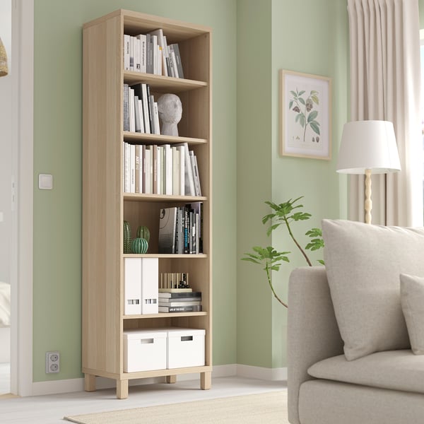 BESTÅ boekenkast in licht eikenhout met boeken en decor tegen groene muur. Beige fauteuil en lamp in de buurt.