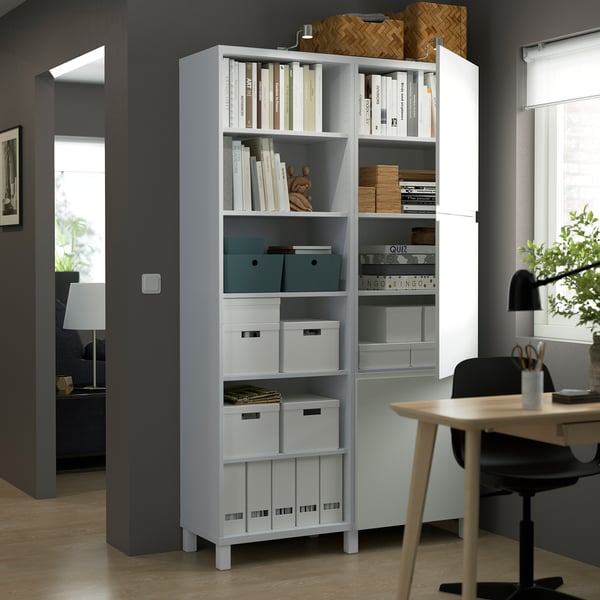 Wit BESTÅ opbergmeubel met verstelbare planken, met boeken en dozen, naast een houten bureau en een zwarte stoel in een thuiskantooromgeving.