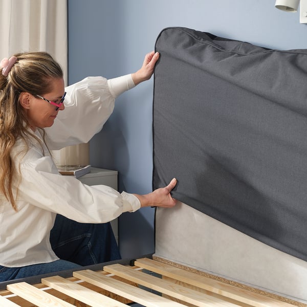Een vrouw bevestigt een donkergrijs hoofdeinde aan een lichtgekleurd houten bedframe en zorgt ervoor dat het veilig en veilig gebruik is.