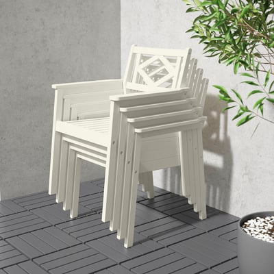 Stapel van drie witte acaciaterrasstoelen met armleuningen op grijze dektegels in de buurt van een muur en plant.