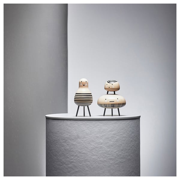 Twee houten figuren met gestreepte patronen op ronde sokkels tegen een grijze muur. De ene figuur heeft een lachend gezicht, de andere een neutrale uitdrukking.