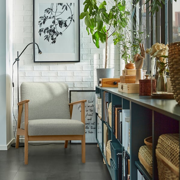 Grijze EKENÄSET fauteuil tegen witte muur met boekenplank, lamp en plant.