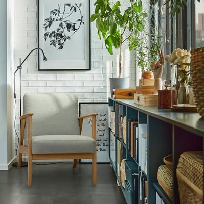 Grijze EKENÄSET fauteuil tegen witte muur met boekenplank, lamp en plant.