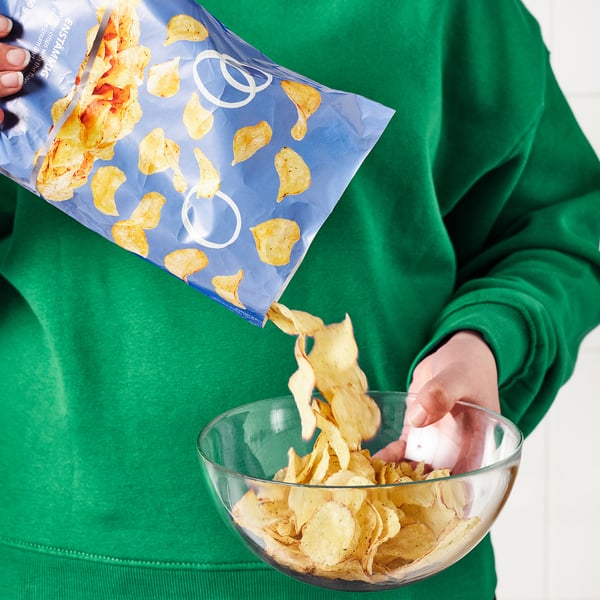 Persoon die chips in een glazen kom giet uit een blauw pakje met het label ENSTÄMMIG terwijl hij een groen sweatshirt draagt.