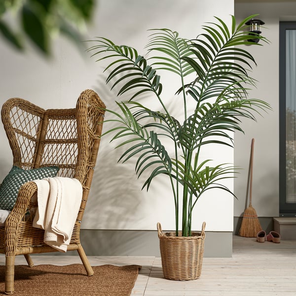 Grote FEJKA kunstplant in een gevlochten mand naast een rotan stoel op de veranda.