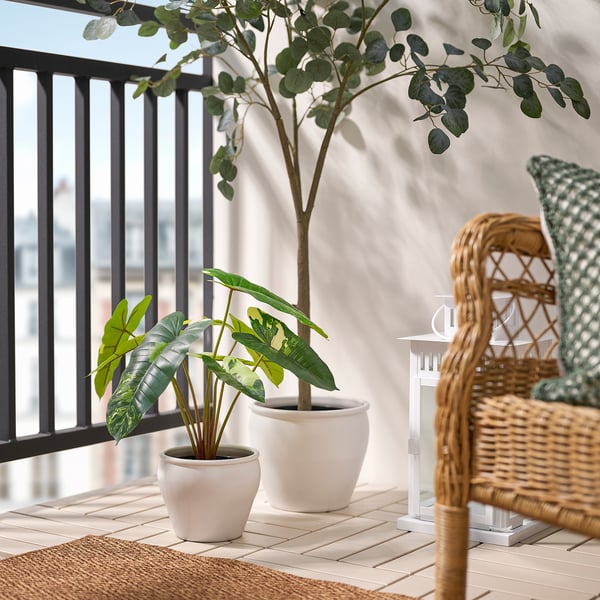 Balkon met FEJKA witte potplanten, een hoog, weelderig, groen; andere klein, levendig. Rieten stoel met kussen rechts, reling links.