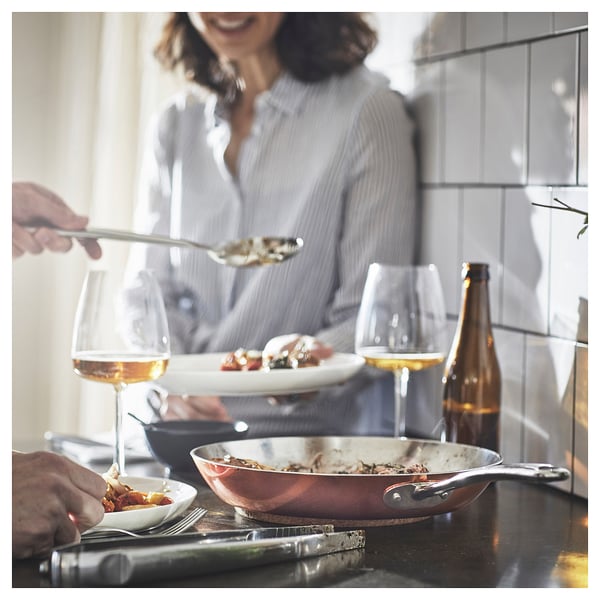 Keuken: persoon die voedsel serveert uit koperen pan op donkere toonbank, wijn, borden.