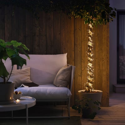 Kleine, verlichte GETKÅL-lichtsnoeren versieren een potplant, omgeven door een gezellig terras met zachte zitplaatsen.