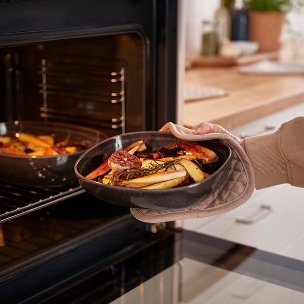 Persoon die een donkergrijze ovenschotel gevuld met geroosterde groenten in een open oven plaatst.