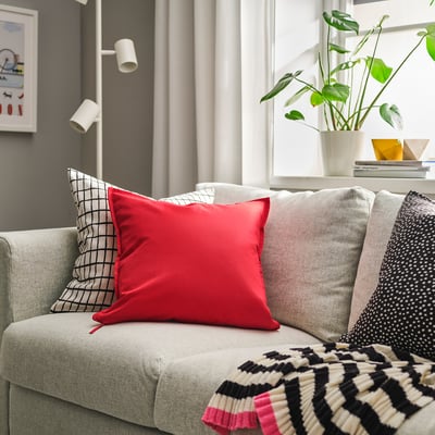 Felrood GURLI-kussen op grijze bank, omgeven door andere kussens, in de buurt van raam met plant.
