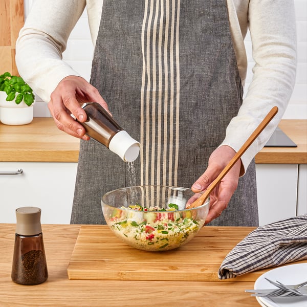 Persoon die salade met pepermolen op houten lijst op smaak brengt. Doorzichtige glazen kom en kruidenpotjes zichtbaar.