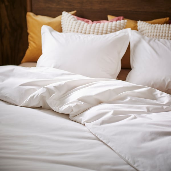 Een fris, wit bed met een luxueus katoenen dekbed en meerdere decoratieve kussens, netjes geplaatst tegen een houten hoofdeinde.