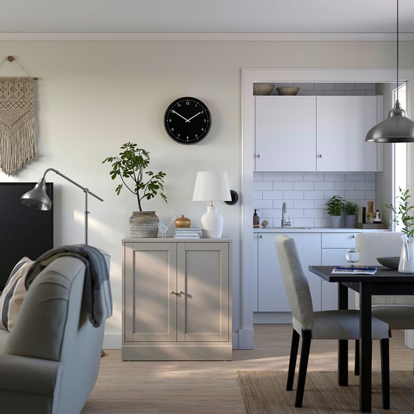 Moderne woonkamer met een rustieke grijze HAVSTA-kast, een zwarte wandklok, stoelen met kussens en houten vloeren.
