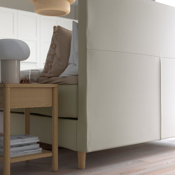 Modern divanbed met een gedempt hoofdeinde voor leesondersteuning, in neutrale tinten, met houten nachtkastje en lamp.