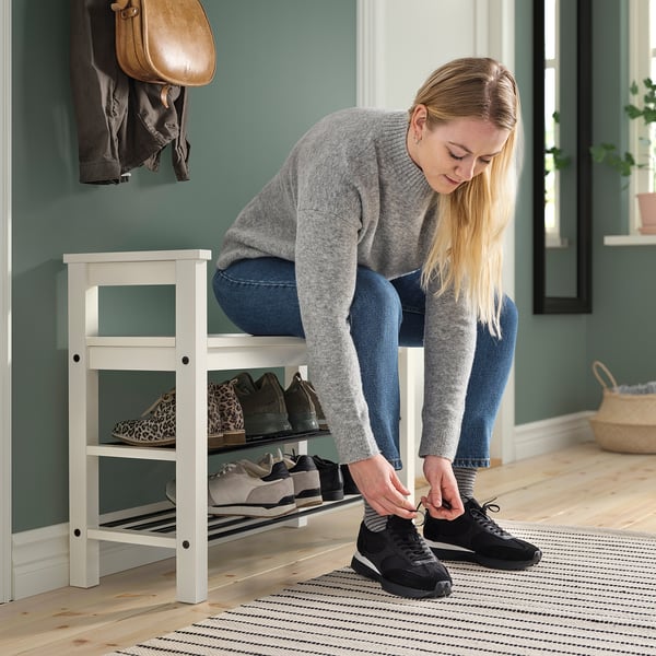Vrouw die zwarte sneakers bindt terwijl ze op een witte HEMNES-bank in de buurt van een schoenenrek zit.