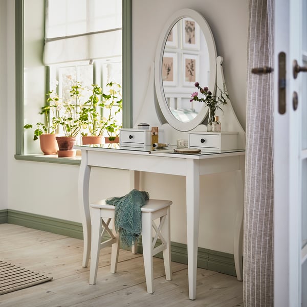 Witte HEMNES kaptafel met spiegel, lades en planten. Houten poten, glazen blad, placée bij raam.