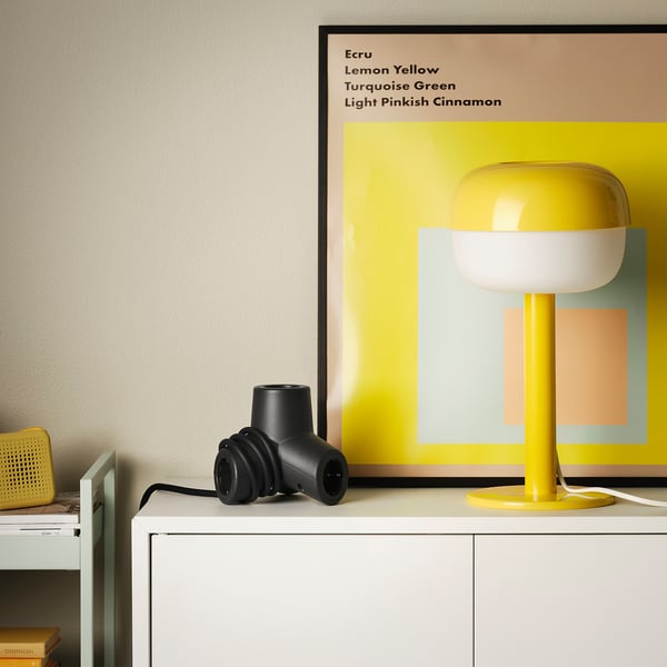 HORNFELS zwarte uitbreidingsdoos, klein, staat op witte meubels, naast gele lamp, naast tv.