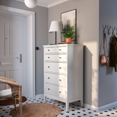 Moderne entree met witte IDANÄS kist, jute tapijt, kapstokken, lamp en planten. Zwart-wit geruite vloer.
