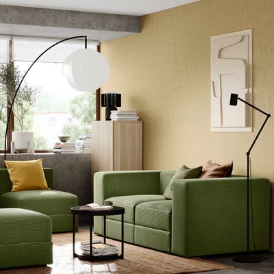 Moderne woonkamer met groen JÄTTEBO bankstel, gele kussens, zwarte vloerlamp en beige kunst aan de muur.