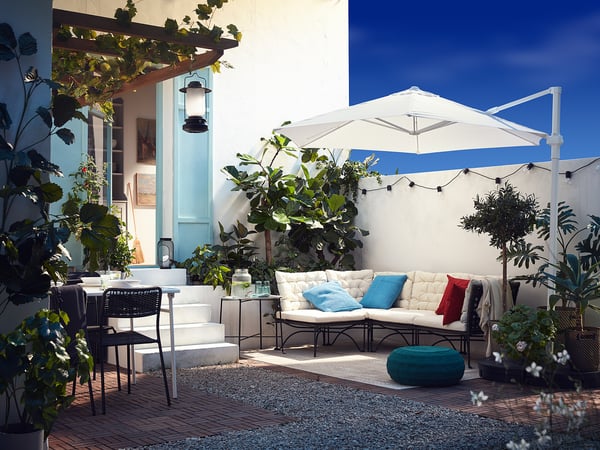 Lichte patio met witte JUTHOLMEN bank, blauwe kussens, planten en witte parasol.