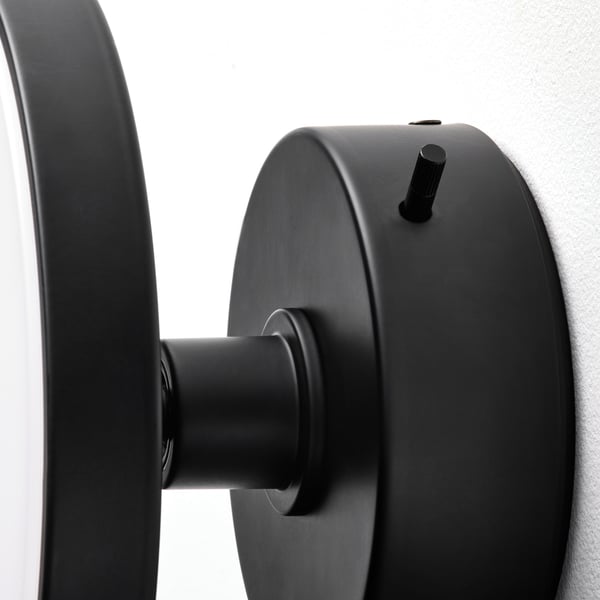 Zwarte wandlamp met verwijderbare spiegel, strak design, gemonteerd op lichtgrijze wand, moderne art deco stijl.
