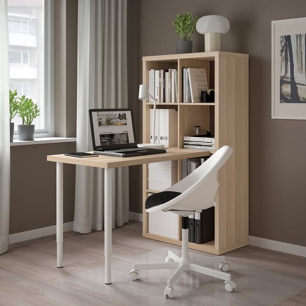 Modern kantoor aan huis met licht houten KALLAX bureau en rek. Witte stoel, lamp en laptop. Grijze muren en groot raam.