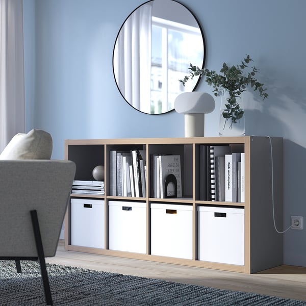 KALLAX boekenkast in lichtgrijs met licht hout, rechthoekig, minimalistisch design. Zit laag, past onder een ronde spiegel, met boeken en een lamp in de hand.