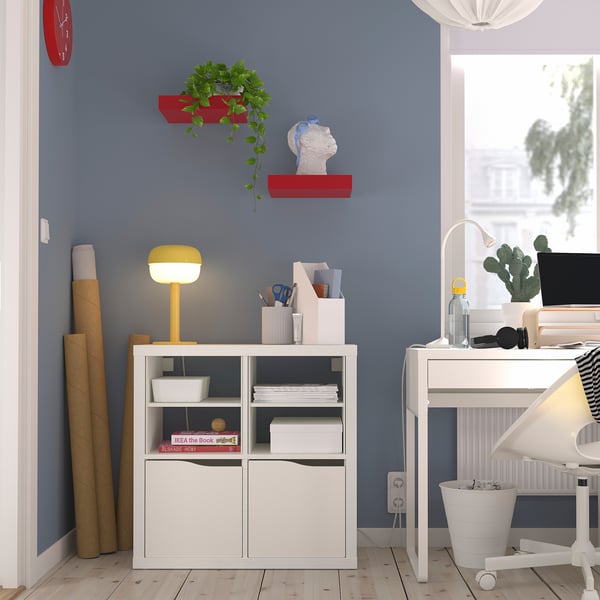 Thuiskantoor met KALLAX-plank, boeken, heldere lamp, rode plantenplanken, klok, bureau, computer. Blauwe muur, houten vloer.
