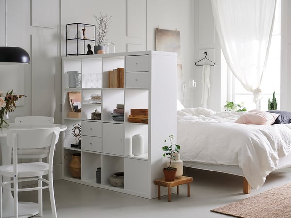 Minimalistische slaapkamer met witte KALLAX planken, met boeken, vazen en decor. Een bed met wit linnengoed en een potplant in de buurt.