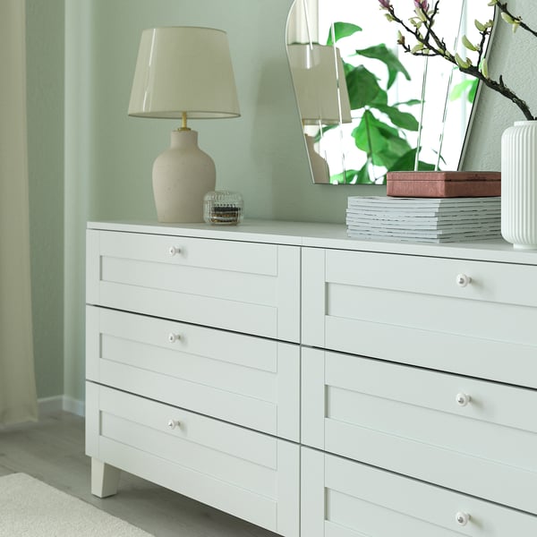 Wit dressoir met zes lades. Decoratieve lamp, spiegel, stapel boeken en vaas. Knoppen zijn wit porseleinachtig.
